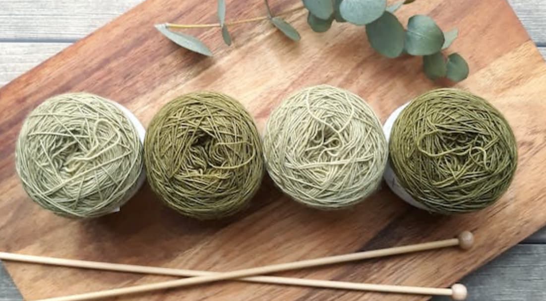 Three balls of green yarn with knitting needles on a wooden board, with eucalyptus leaves in the background