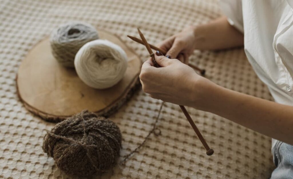 A person's hands knitting with wooden needles, with balls of white and grey yarn and a partially knitted piece on a wooden board