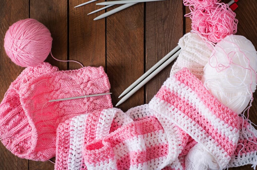 Pink and white knitted garments with yarn balls and needles on wood