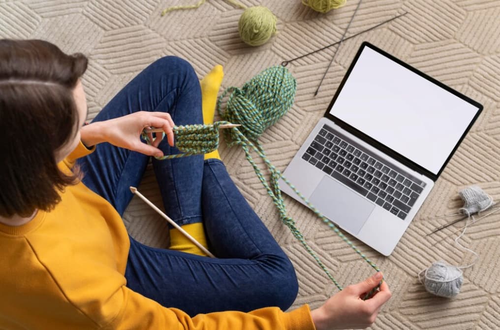 An individual knitting beside an open laptop, crafting a green piece