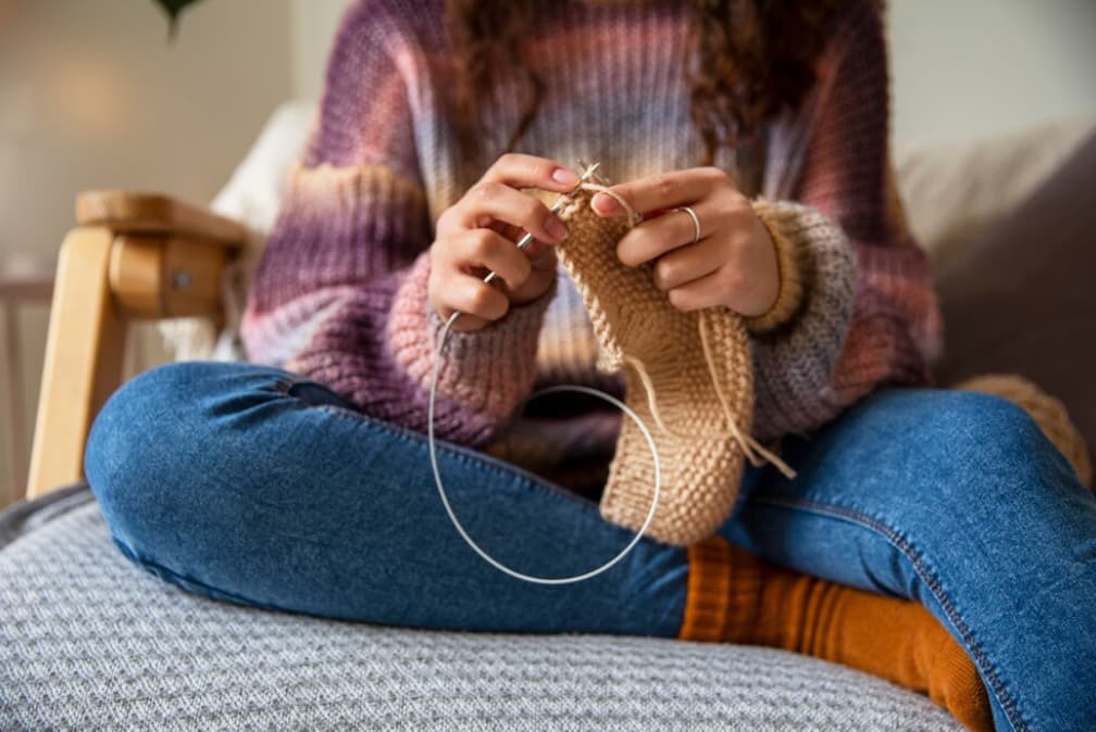 woman in jeans and a colorful sweater knitting the sock and sitting in a chair