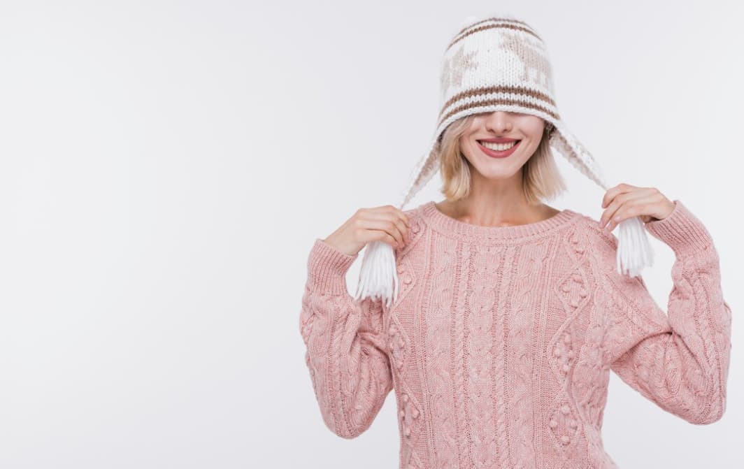 A smiling woman playfully pulls on the tassels of her white patterned winter hat