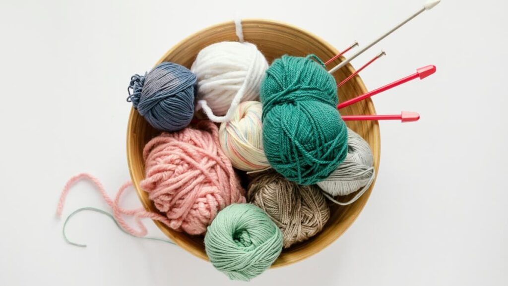 colorful green, blue, gray, white and brown yarns with red needles inside the wooden bowl