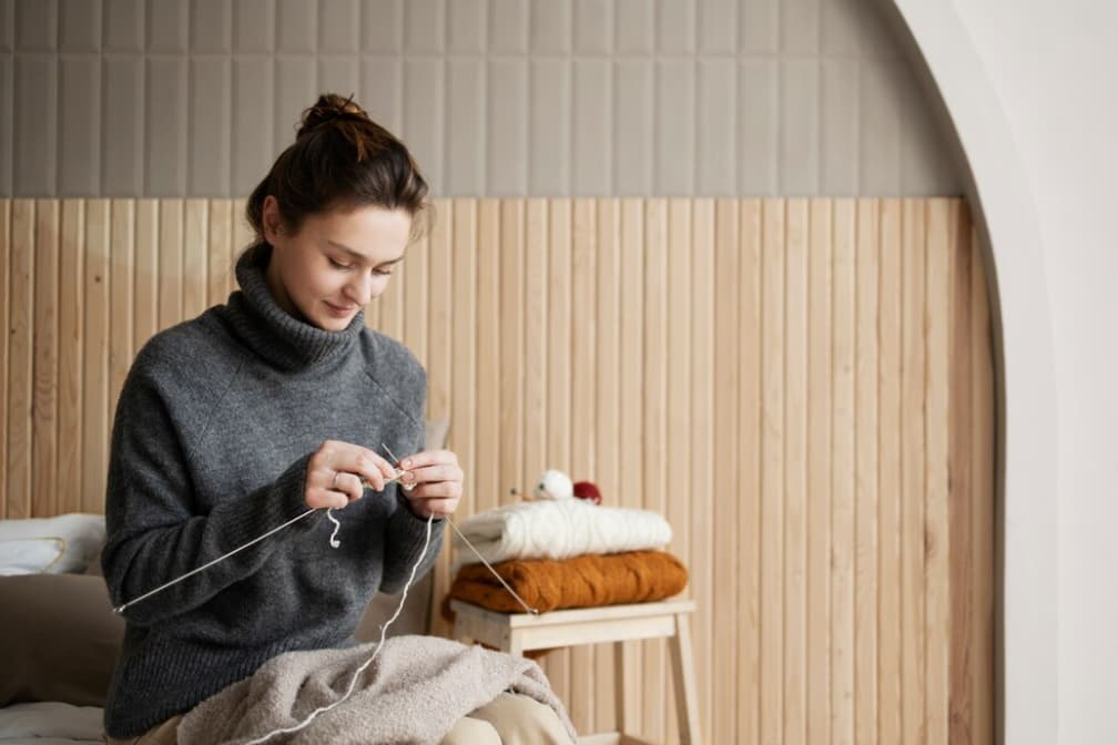 A woman in a gray sweater sits on the bed and knits with needles, the wooden wall behind her