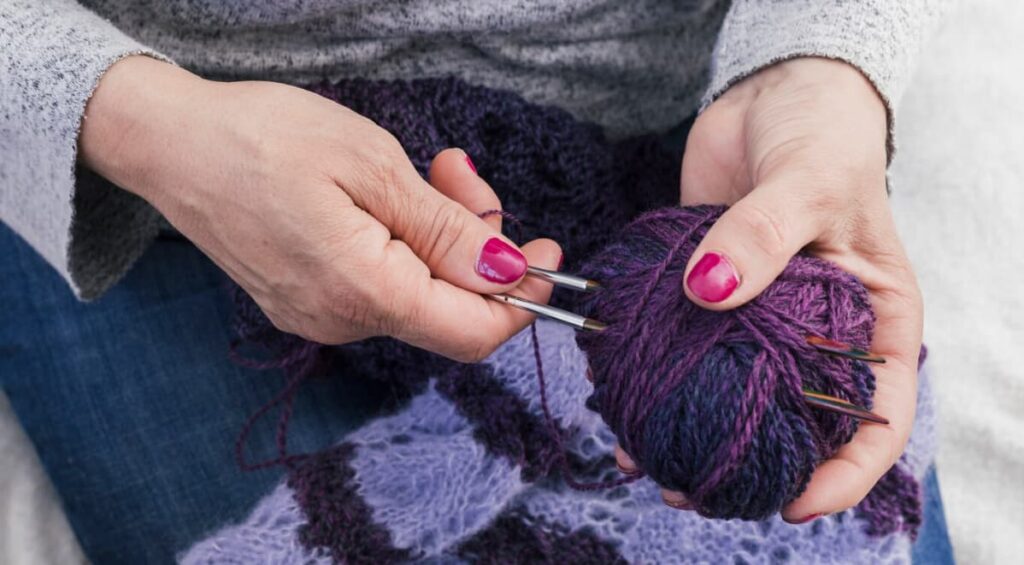 A woman in a gray sweater and jeans holds a purple yarn and needle and knits a scarf