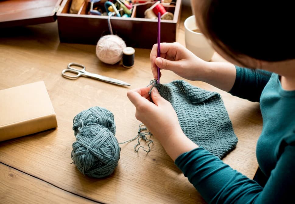 Person crocheting a grey fabric with blue yarn on a wooden table