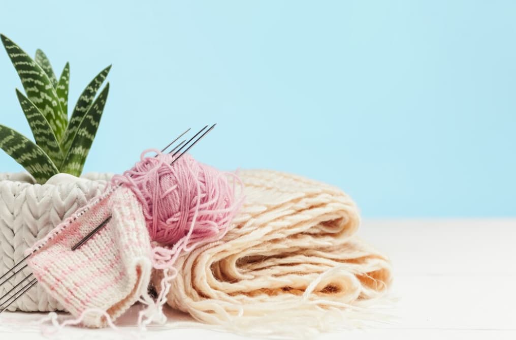 a vase with flowers, a ball of pink wool, and needles on a white wooden table